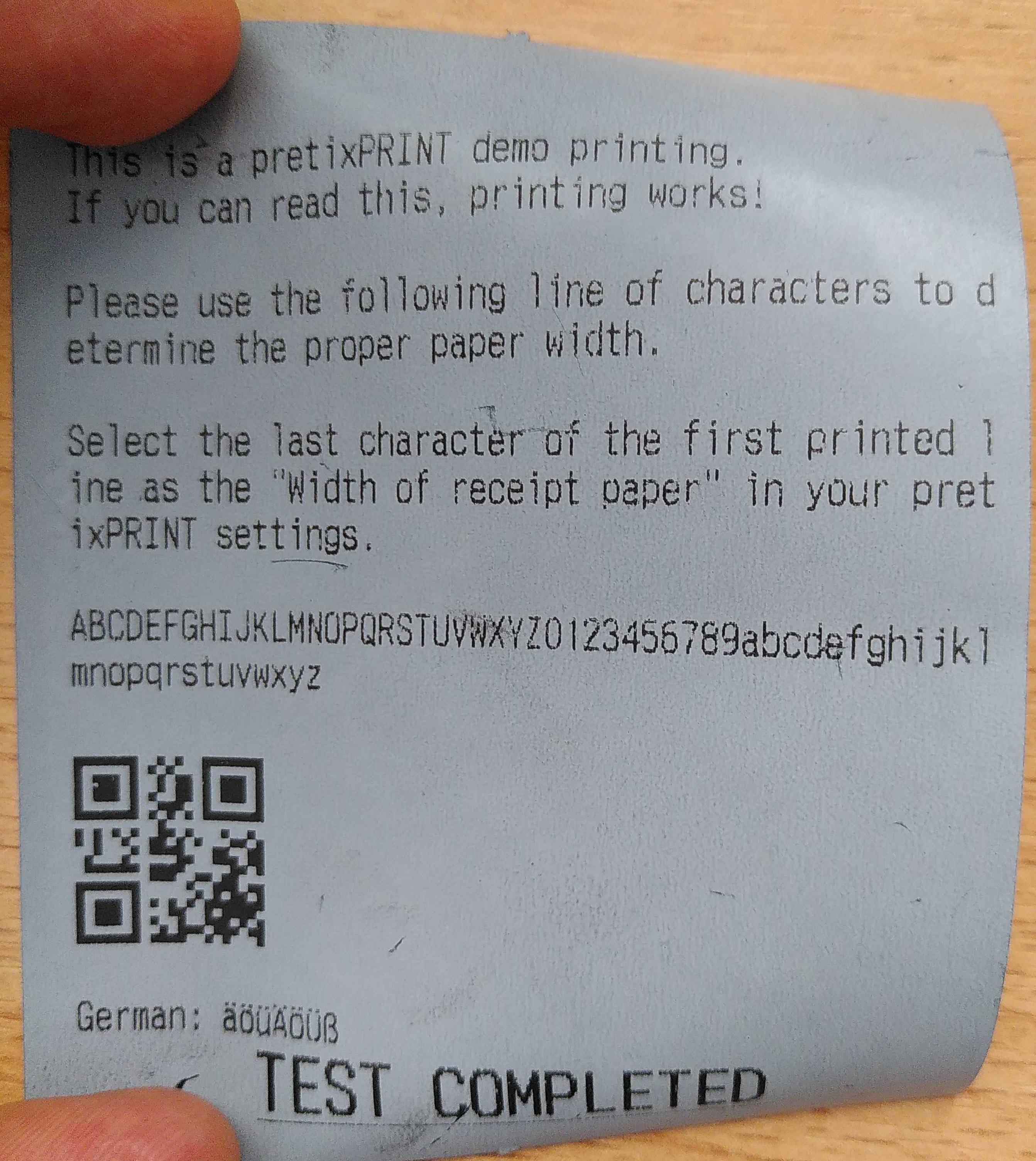"Ein Streifen graublaues Belegpapier mit Text, einer Zeile zum Testen der Breite des Belegpapiers, und einem QR-Code"