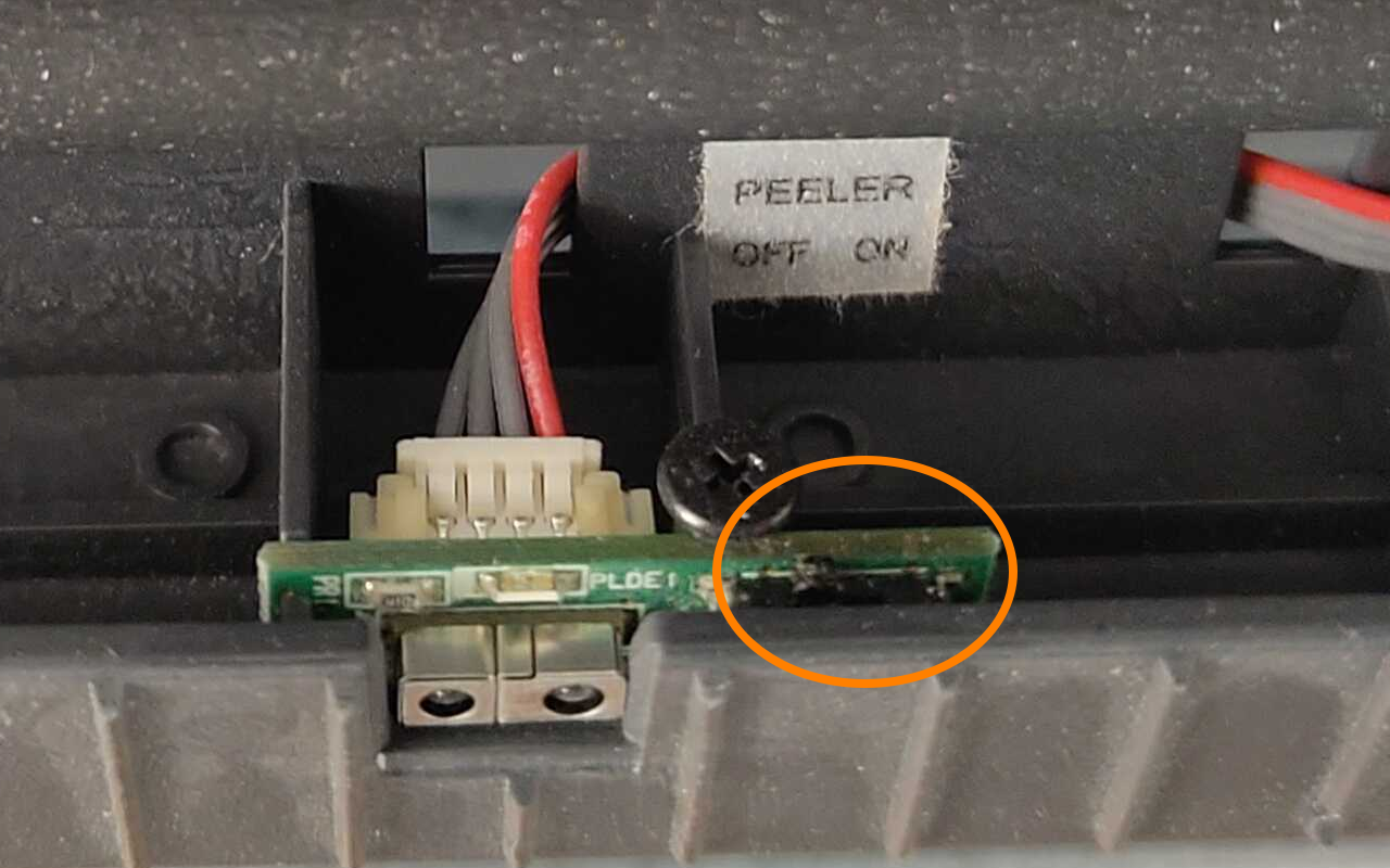 Nahaufnahme des Peeler-Controllers, einer kleinen Platine, von der vier Kabel ins Innere des Geräts laufen. Ein Etikett oberhalb der Platine zeigt den Text "PEELER OFF ON". Ein orangefarbenes Oval markiert den Umschalter auf der Platine
